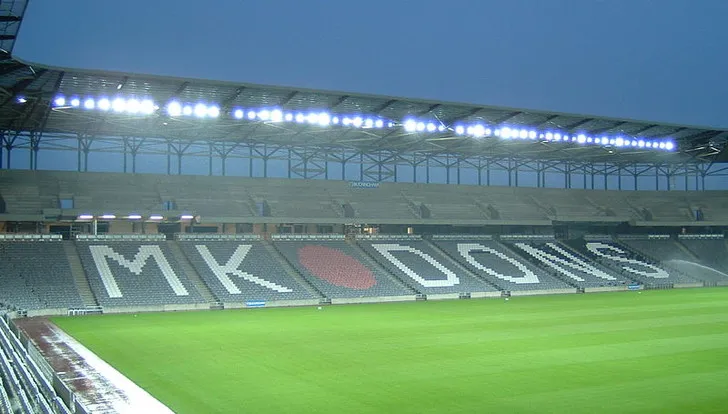 Một góc khán đài sân vận động MK Dons với hàng ghế trắng đỏ và logo câu lạc bộ, thể hiện sự cổ vũ của người hâm mộ bóng đá Anh.
